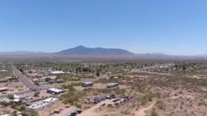 Aerial view of Bisbee, AZ, reveals a small town with scattered buildings and streets in a desert landscape. In the background, a mountain range rises under a clear blue sky. Sparse vegetation dots the dry terrain, and cars are parked in an RV park. The scene captures affordable living in a quiet, expansive setting.