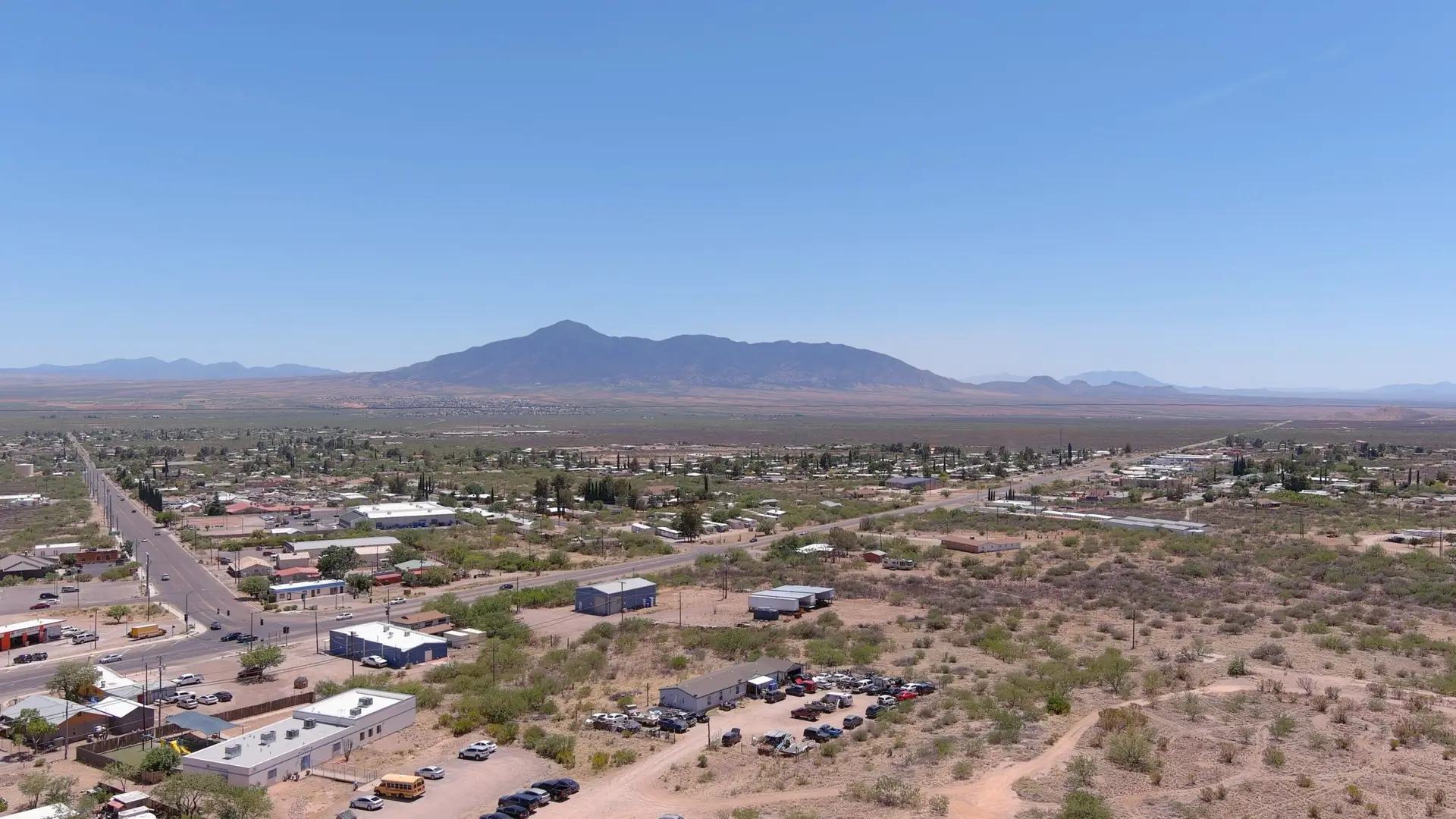 Aerial view of Bisbee, AZ, reveals a small town with scattered buildings and streets in a desert landscape. In the background, a mountain range rises under a clear blue sky. Sparse vegetation dots the dry terrain, and cars are parked in an RV park. The scene captures affordable living in a quiet, expansive setting.