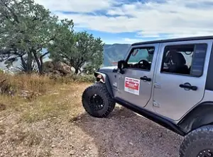 A silver off-road vehicle with large tires is parked on a rocky dirt path, leading into Bisbee, AZ. The path is flanked by lush green bushes and trees, with a sign visible on the vehicle's door. Mountains rise in the background under a partly cloudy blue sky.
