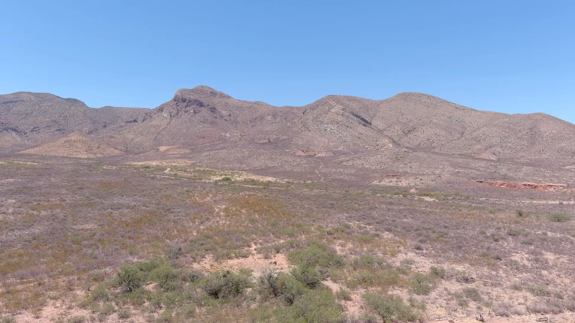 A wide, barren landscape with sparse green shrubs covers the foreground near Bisbee, AZ. The background features rugged, brown desert mountains under a clear blue sky, creating a stark contrast between the earthy tones and the sky above. The terrain looks dry and rocky, typical of a desert environment.