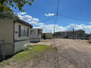 A sunny mobile home park offers affordable living with three homes along a dirt road in Bisbee, AZ. In the foreground, a white trailer sits behind a chain link fence, while two others are visible in the background. Overhead, scattered clouds mingle with power lines against a clear blue sky.