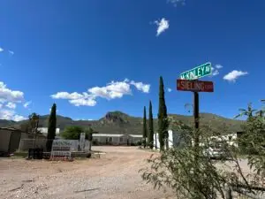 A rural scene with a dirt road intersection showcases street signs for McKinley Ave and Sieling Loop. In the background, mountains rise under a clear blue sky. Several tall cypress trees, a Parc Ridge sign, an RV Park nearby, a parked car, and a mobile home are also present.