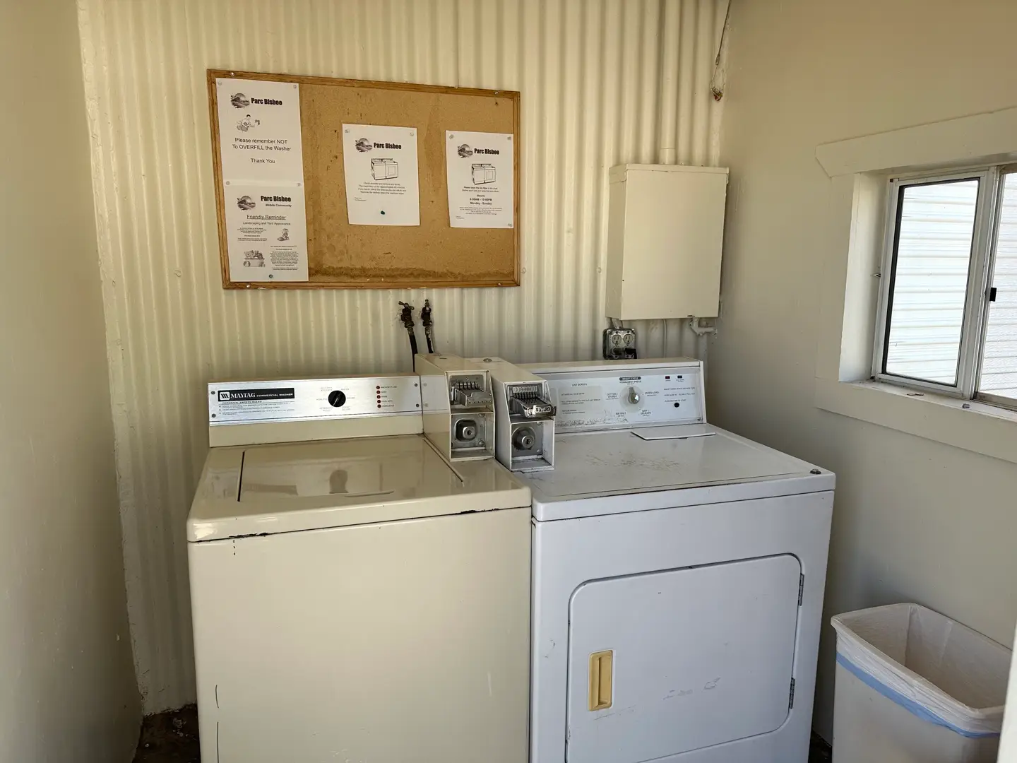 A small laundry room at the RV park features a white top-loading washing machine and a matching dryer. Above them, a cork bulletin board displays instructional flyers. The room has corrugated walls, a small window, and a white trash bin in the corner, offering affordable living amenities.