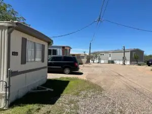 A mobile home park near me features two visible homes under a clear blue sky, with an SUV parked on the gravel driveway. A utility pole with overhead cables crosses the scene, while grass and gravel lay in the foreground, complemented by sparse vegetation typical of Bisbee, AZ.