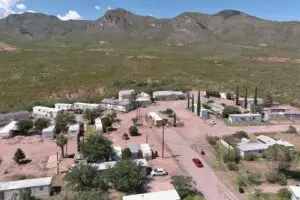Aerial view of a small, affordable community in Bisbee, AZ, with scattered homes and trailers surrounded by greenery. Two cars are parked on a dirt road near an RV park. Tall cypress trees are interspersed among the residences. Mountains rise in the background under a partly cloudy blue sky.