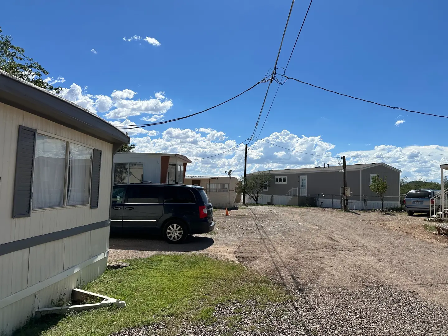 A sunny day at a mobile home park with a clear blue sky and scattered clouds. Several trailers line the gravel road, while a black SUV sits parked beside one. Utility wires stretch overhead, framing small patches of grass and rocky terrain in the area.