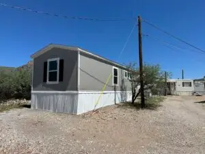 A single-story mobile home with gray siding and white trim sits under a clear blue sky in an affordable living community. It features two windows with dark shutters, and a dirt path leads to the home, flanked by utility poles and sparse vegetation. Another mobile home is visible in the background.