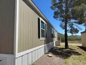 A beige mobile home with white trim stands in a serene Mobile Home Park under a clear blue sky. It features two windows with black shutters, a large tree to the right, and views of distant hills near Bisbee, AZ. The dry ground has sparse grass and gravel, reflecting affordable living in this picturesque area.