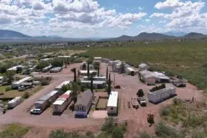 Aerial view of a rural landscape featuring a mobile home park arranged in a semi-circle, embodying affordable living. The area is surrounded by green vegetation and distant mountains under a partly cloudy sky. Parked vehicles and construction machinery are visible on the dirt roads.