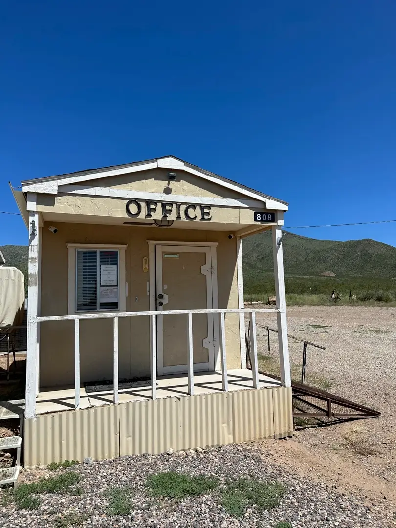 Small beige building labeled OFFICE, perfect for RV Park visitors. A white railing and steps lead to the door, with a window displaying papers next to it. The building sits on a gravel area with grass and mountains under a clear blue sky. Number 808 is displayed prominently, offering affordable living solutions nearby.