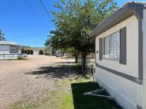 A small, affordable mobile home park with several homes basks in the sun. A white mobile home with closed shutters sits in the foreground on the right. Gravel covers the ground, while a large tree offers shade. In the background, a clear blue sky and distant hills complete this tranquil scene.