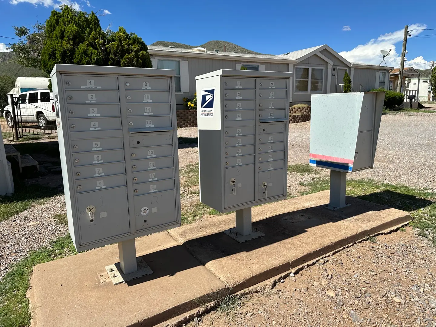 A cluster of metal mailboxes stands on a concrete slab in a rural area near Bisbee, AZ. Two large units with multiple compartments accompany a smaller box with red and blue stripes. In the background are trees, a mobile home, mountains, and a white van parked under a clear blue sky.