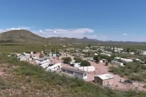 Aerial view of a small rural settlement near Bisbee, AZ, surrounded by desert landscape and mountains under a clear blue sky. The area features scattered houses, trailers, and RV parks amid patches of greenery. Multiple dirt roads meander through the settlement, with distant mountain ranges adding depth to the scene.