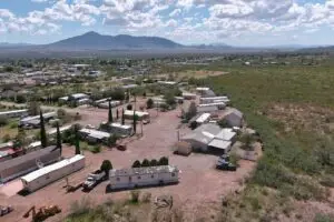 Aerial view of a rural residential area with scattered houses and trailers amidst greenery. This mobile home park embraces affordable living as a dirt road winds through the community. Mountains rise in the background under a cloudy sky, with several vehicles visible near the homes.
