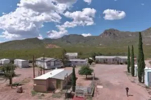A rural trailer park under a bright blue sky with scattered clouds offers affordable living options. Several mobile homes are set on a gravel area with sparse vegetation. Tall trees line a dirt road as someone walks nearby, and the scenic mountains of Bisbee, AZ rise in the background.