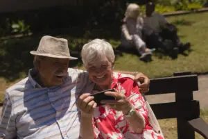 An elderly couple sits on a bench in Bisbee, AZ, smiling and looking at a smartphone. The man wears a beige hat and striped shirt, while the woman dons a red floral blouse. In the background, another couple relaxes on the grass. It's an affordable living paradise under bright sunshine with lush greenery around.