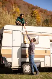 A woman sits on top of a vintage beige and brown camper van in Bisbee, AZ, reaching down to a man beside it. She wears a green and black plaid shirt, while he sports plaid and jeans. They smile against a backdrop of autumnal trees on a sunny day, celebrating the essence of RV park adventures.