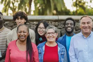A diverse group of seven smiling adults stand outdoors in front of a stone wall and greenery, embodying the vibrant community spirit of Bisbee, AZ. They vary in age and ethnicity, showcasing affordable living with their casual, colorful clothing amidst blurred trees and sunlight.