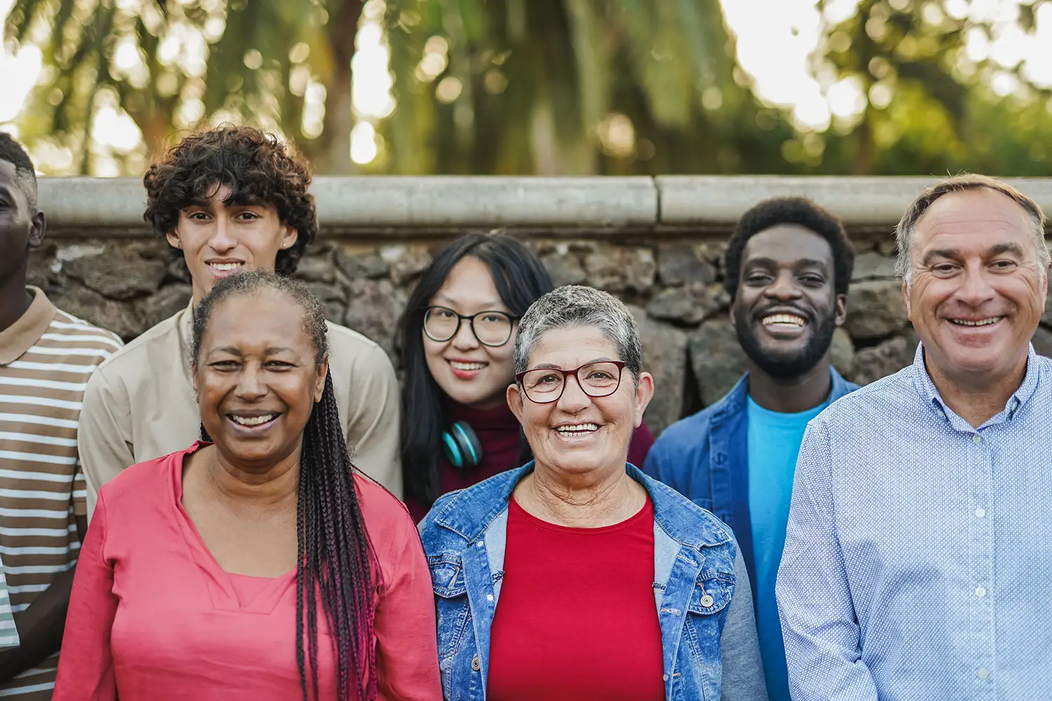 A diverse group of seven smiling adults stand outdoors in front of a stone wall and greenery, embodying the vibrant community spirit of Bisbee, AZ. They vary in age and ethnicity, showcasing affordable living with their casual, colorful clothing amidst blurred trees and sunlight.