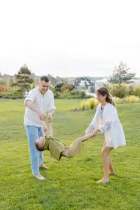 A man and woman joyfully swing a child by the arms and legs in a lush green park near Bisbee, AZ. The child wears a khaki outfit, the man is in a white shirt and jeans, and the woman dons a white dress with blue embroidery. Trees and a clear sky create the perfect backdrop for this lively family moment.