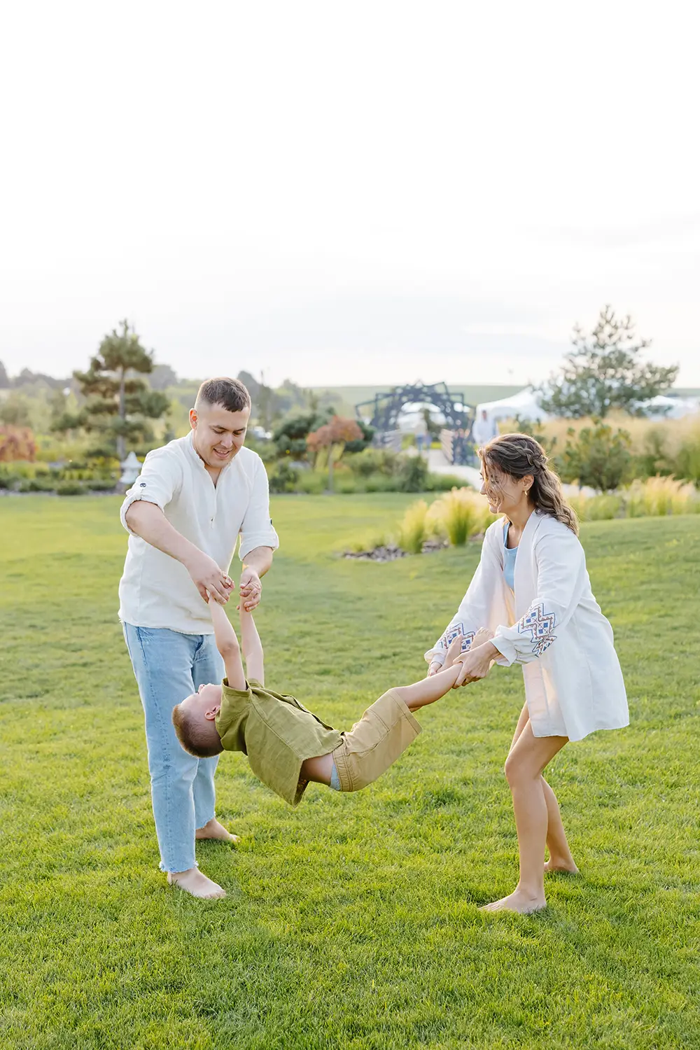 A man and woman joyfully swing a child by the arms and legs in a lush green park near Bisbee, AZ. The child wears a khaki outfit, the man is in a white shirt and jeans, and the woman dons a white dress with blue embroidery. Trees and a clear sky create the perfect backdrop for this lively family moment.