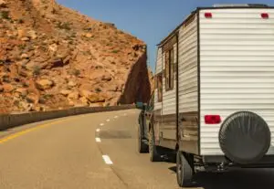 A pickup truck tows a white camper trailer along a winding road through a rocky desert landscape, reminiscent of the picturesque surroundings near Bisbee, AZ. The clear blue sky contrasts with reddish-brown cliffs as the road curves gently left, evoking a journey toward an affordable living oasis.