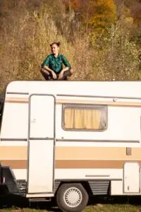 A person in a green plaid shirt and black pants is sitting on the roof of a beige and brown camper, parked at an RV Park near Bisbee, AZ. Surrounded by grassy fields and autumn trees with vibrant yellow and orange foliage, the scene is beautifully lit by natural sunlight.