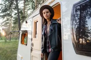 A woman in a black leather jacket and hat stands at the open door of a white camper van, nestled in an RV park. She is surrounded by tall trees and hanging lights. The warmly lit interior of the camper exudes affordable living, while she gazes forward, creating a serene and adventurous atmosphere.