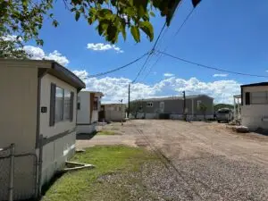 A gravel road winds through Parc Bisbee, a mobile home park in Bisbee, AZ, beneath a bright blue sky dotted with fluffy clouds. Mobile homes line both sides, set against green grass patches and trees. Overhead power lines crisscross the scene, while a distant traffic cone marks the RV hook-up sites.