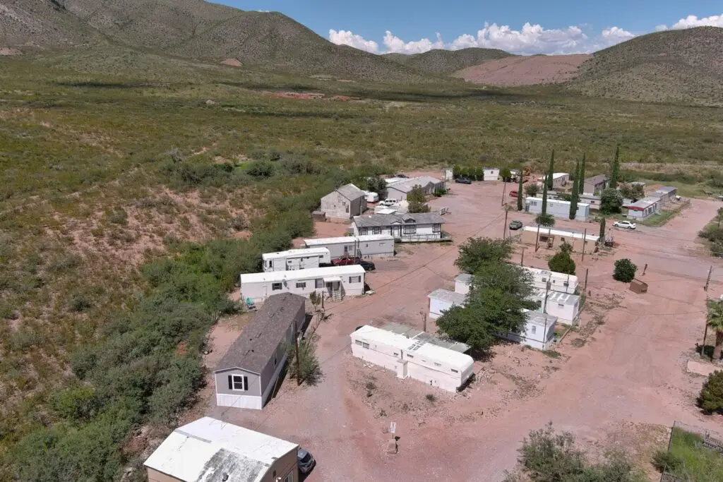 Aerial view of a desert landscape with a small trailer park near Bisbee, AZ. Several white and beige mobile homes are scattered among sparse greenery. Dirt roads connect the homes. The background includes rolling hills and a bright blue sky with fluffy clouds, reminiscent of Parc Bisbee’s charm.